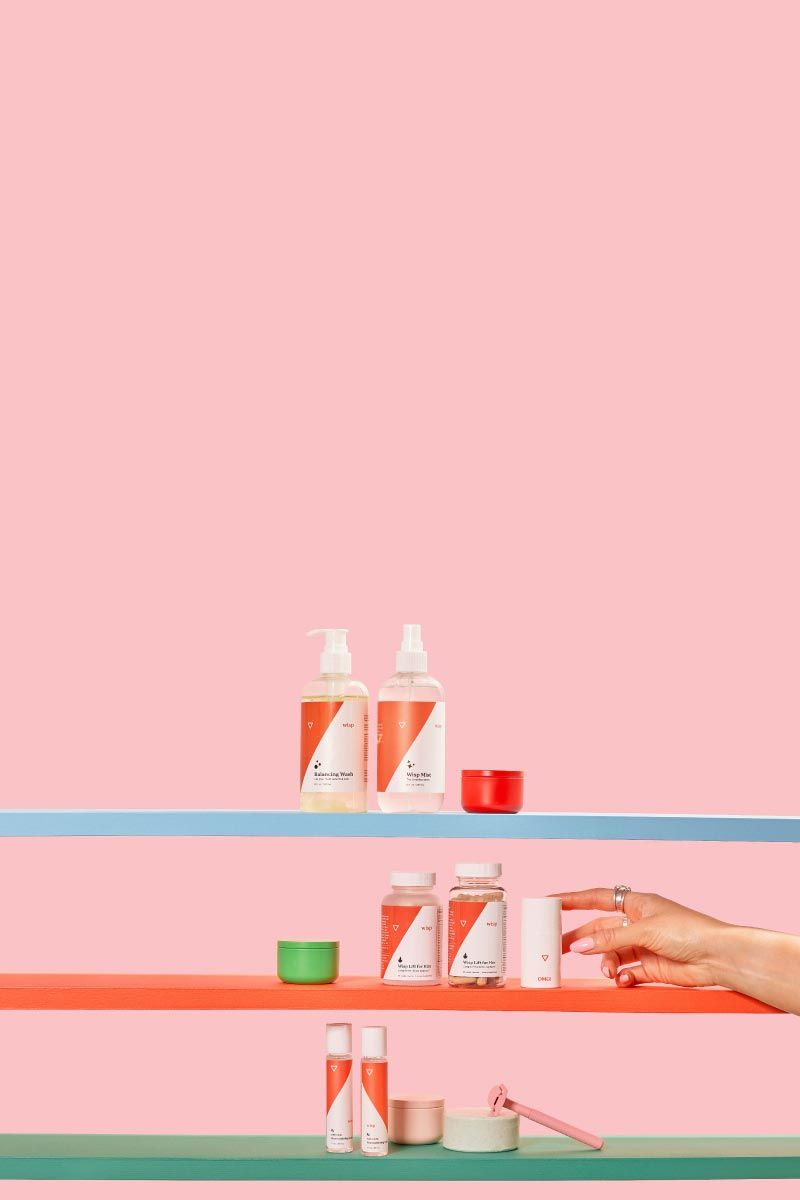 A variety of Wisp products on 3 colorful shelves with miscellaneous containers, a pink razor and a woman's hand reaching for Wisp OMG Cream in front of a pink background