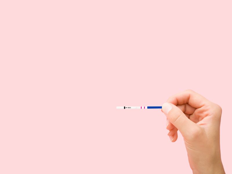 A person's hand holding a testing strip in front of a pink background