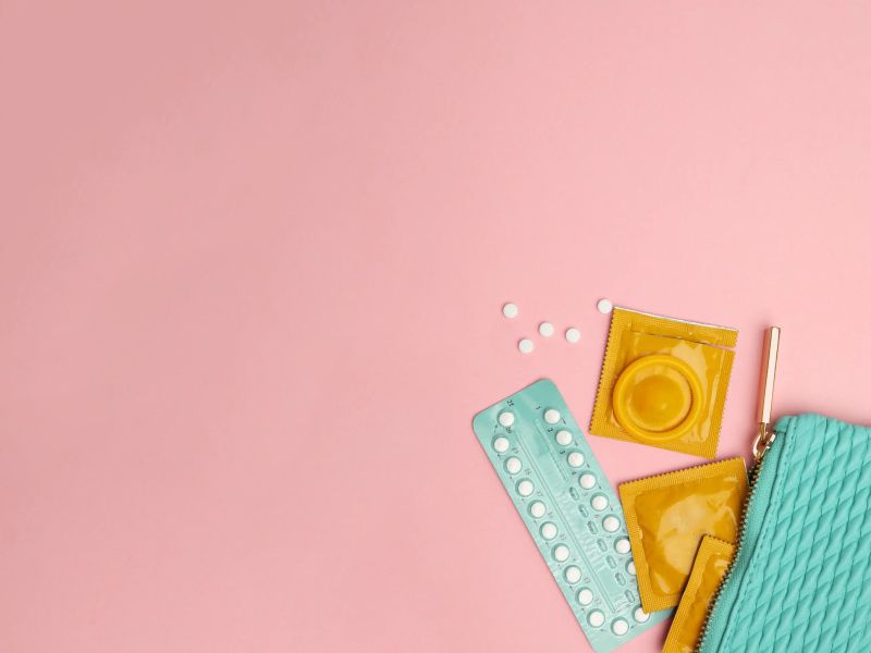 A green pouch with condoms and birth control pills spilling out on a pink surface