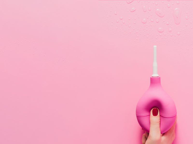 A woman's hand with red fingernails holding a pink vaginal douche bulb over a pink background