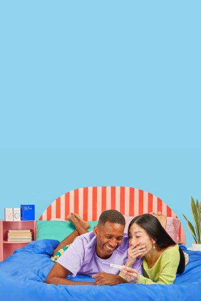 Couple on bed holding pregnancy test looking surprised and excited with Proov products on a pink nightstand and a plant in the corner