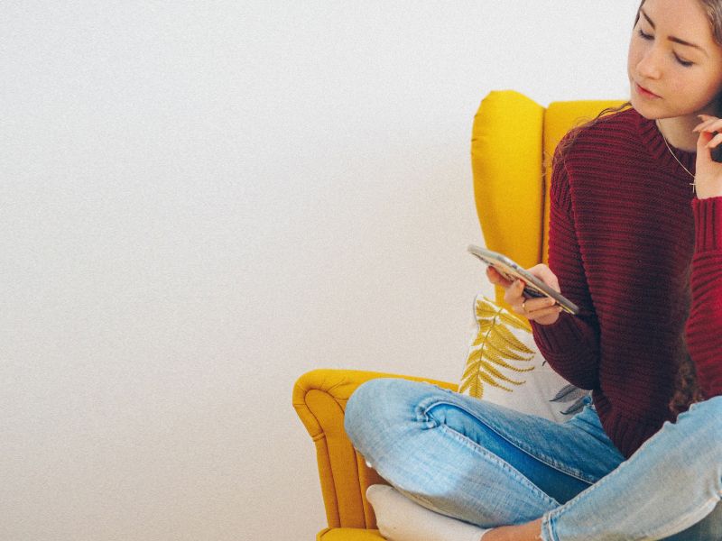 A female wearing a maroon sweater and blue jeans looking up information on her phone while sitting on a yellow chair