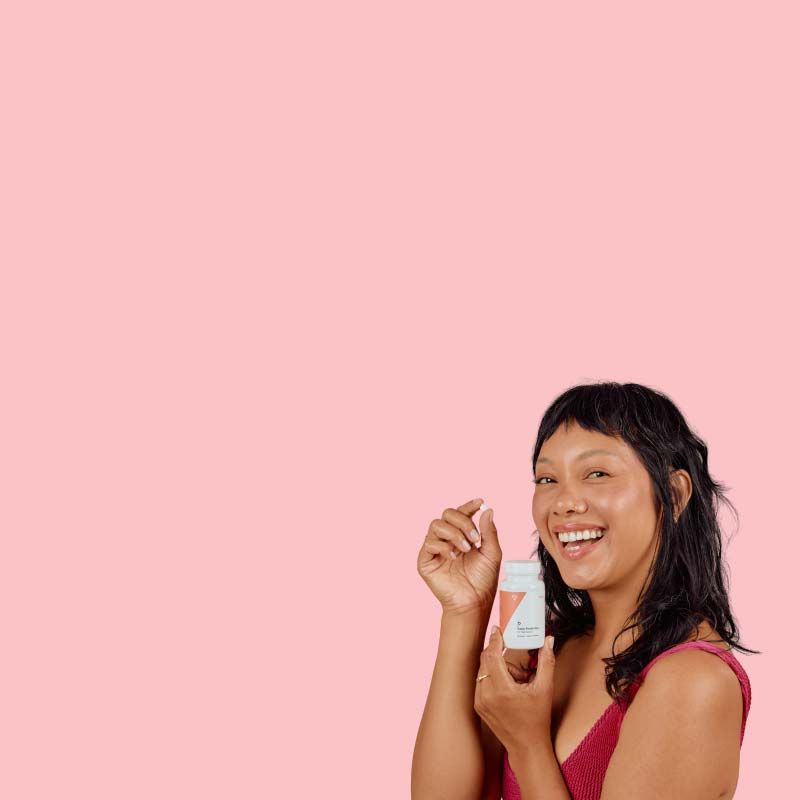 A happy woman wearing a red tank topis holding a pill and a Wisp bottle in front of a pink background