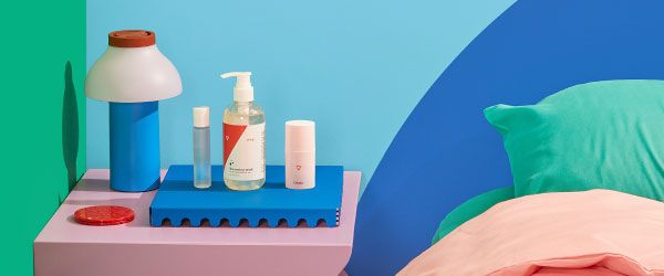 3 Wisp products on a pink nightstand with a blue lamp and next to a bed with colorful bedding and a woman's intertwined legs