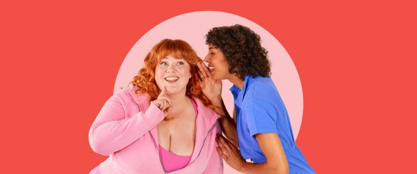 A woman wearing blue is whispering into another woman's ear who is smiling and wearing pink clothing