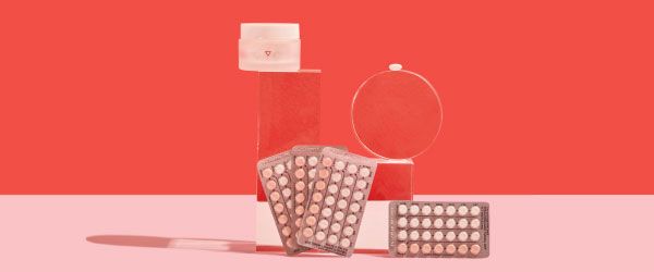 4 birth control packets and a small glass pill jar are balanced on clear abstract shapes on a pink surface with a red background