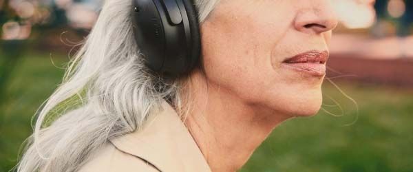An older woman wearing headphones and a trench coat ouotdoors