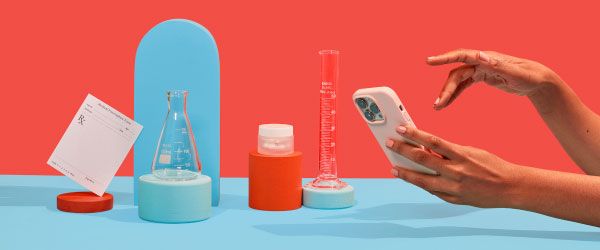 Wisp glass pill jar, test tubes, and a prescription script are balanced on colorful abstract shapes on a blue surface with a red background