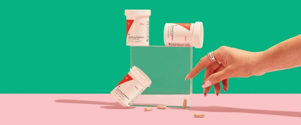 3 bottles of Wisp Equalizing Probiotics balanced on a clear block with pills spilling out and a woman's hand reaching for them on a pink surface with a green background