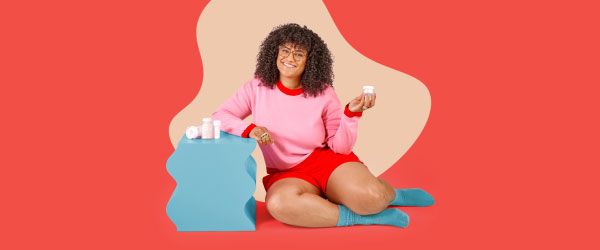 A woman is smiling and wearing a pink and read sweater and shorts with blue socks while sitting on a red surface next to a blue nightstand with several Wisp medications on it