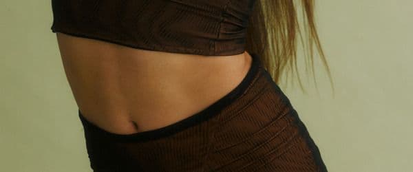 Close-up of a person’s midriff in a brown textured crop top and matching low-rise pants, against a sage green background.
