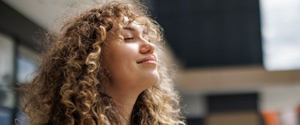 A happy, peaceful woman with curly hair has her head turned up to the sky with sunlight on her face