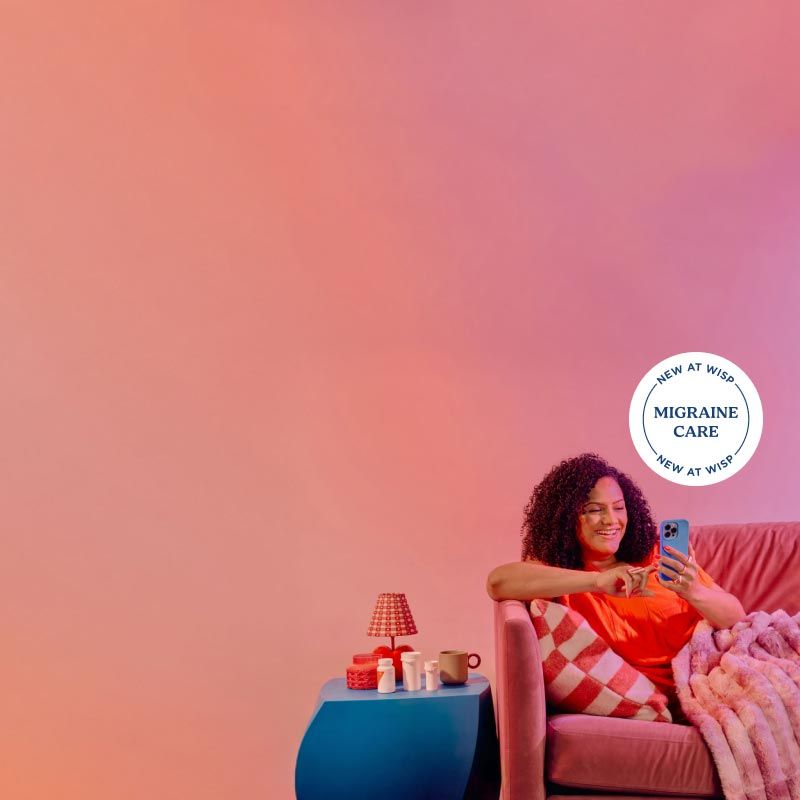 A woman is smiling while using a mobile phone on a couch with a cozy blanket next to a blue nightstand with Wisp medications on it