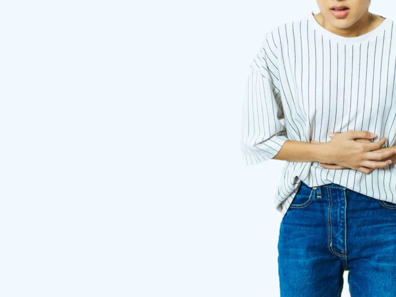 A woman wearing a striped white and black shirt with blue jeans has her hands over her abdomen in obvious discomfort