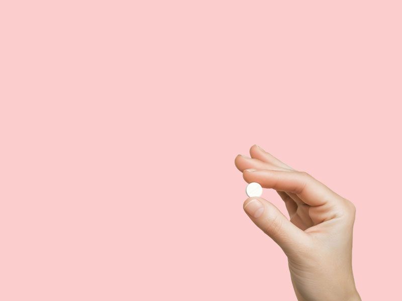 A person's hand holding a small round white pill in front of a pink background