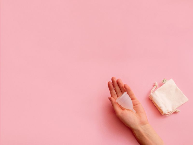 A woman's hand holding a menstrual cup with a storage bag next to it on a pink surface