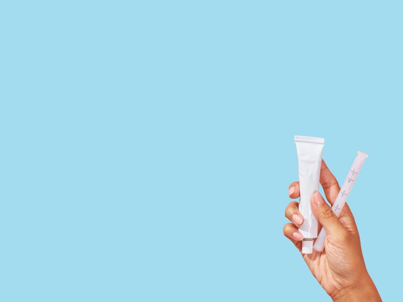 A person's hand is holding a tube of hemorrhoid cream in front of a light blue background