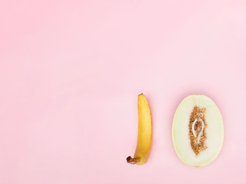 A banana and a cut open vulva-shapes fruit on a pink surface