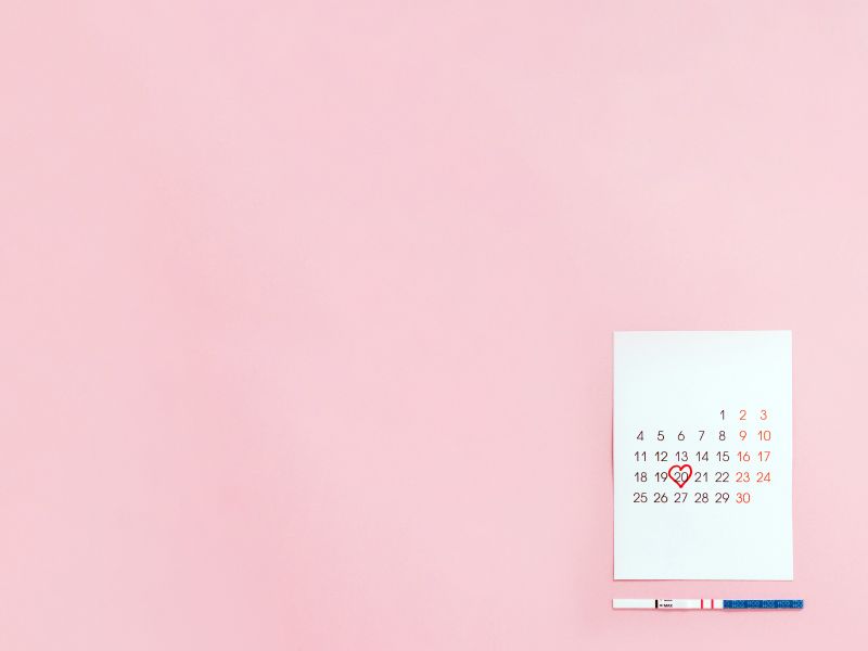 A calendar on a pink surface with a circled date and an ovulation test