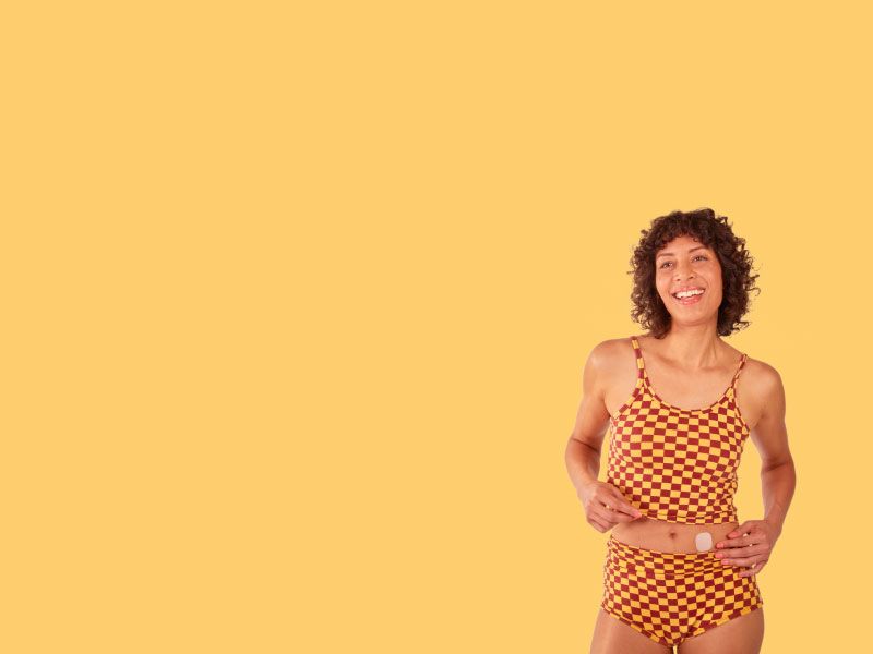 A woman smiling and wearing a checkered yellow and brown tank top and shorts is placing a hormone replacement therapy patch on her abdomen