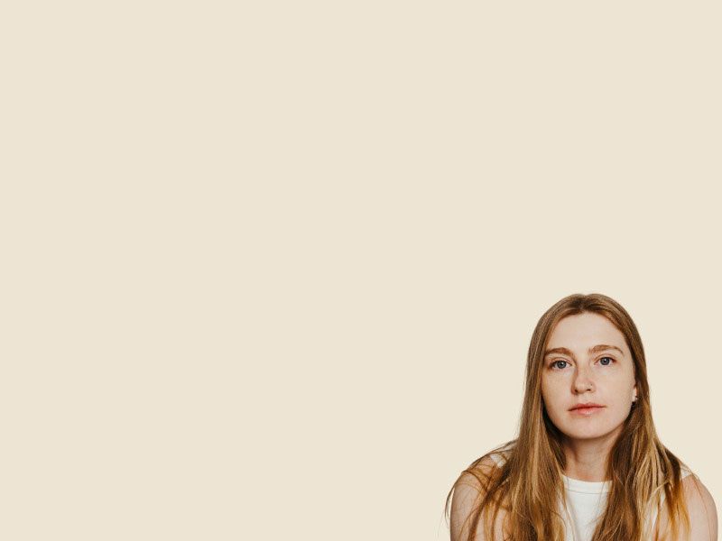 A woman with blonde hair wearing a white tank top looks questioningly at the viewer