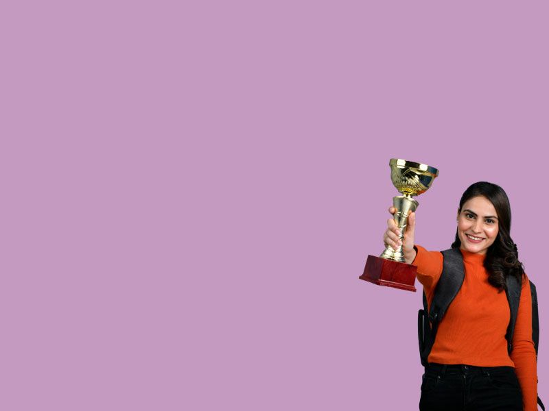 A woman wearing a red sweater, black pants and a black backpack holding a gold trophy in the air in front of a purple background