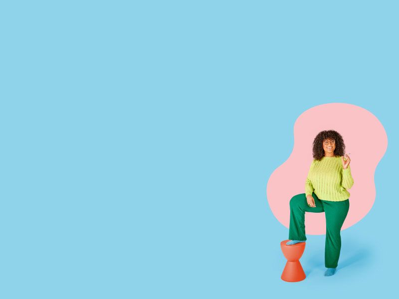 A woman wearing a lime green sweater, dark green pants and light blue socks is smiling and holding a syringe with her foot on on an orange stool