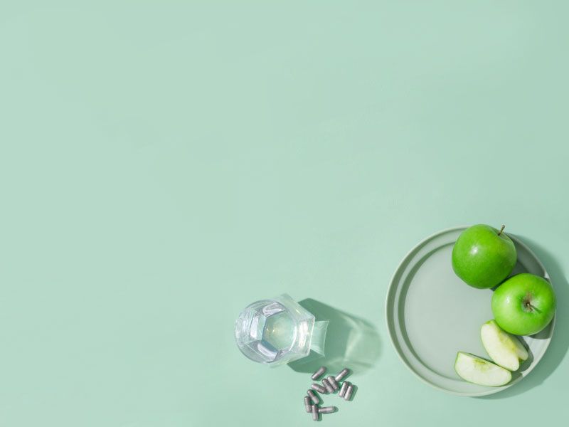 Sliced and whole apples on a plate with supplement capsules next to a glass of water on a green surface