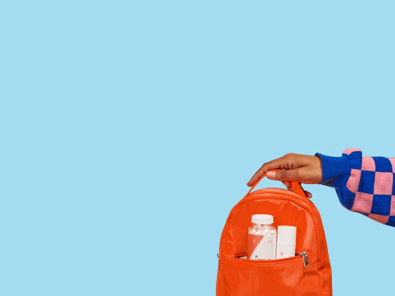 A person's hand holding an orange backpack with Wisp medication bottles in the pocket in front of a light blue background