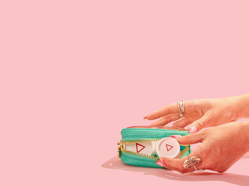 A woman's hand reaching for Wisp pill vials inside a green travel pouch laying on a pink surface