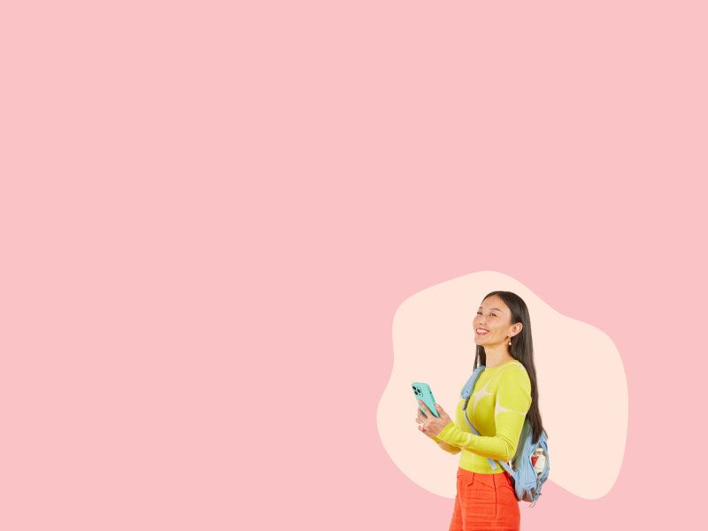 A woman wearing a yellow sweater and orange pants with a light blue backpack containing Wisp medication is smiling and holding a cell phone in front of a pink background