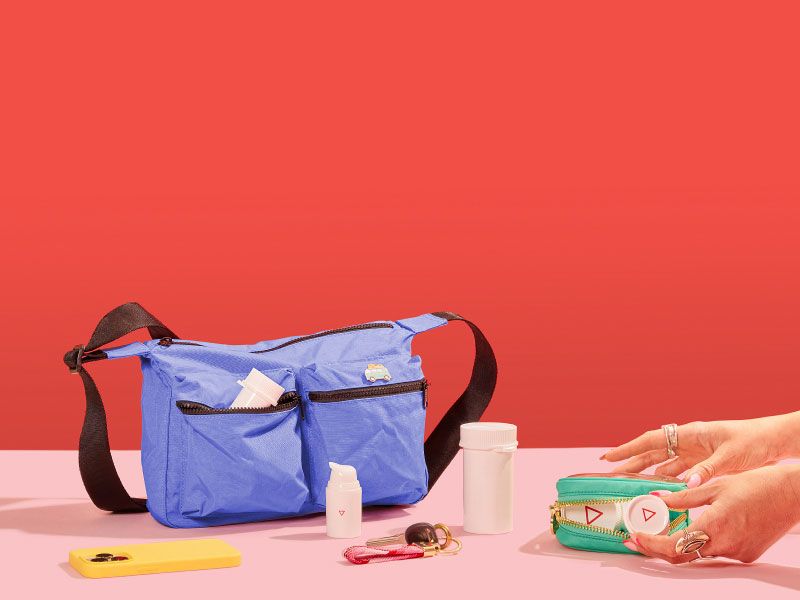 A woman's hands grabbing Wisp medication vials from a travel pouch sitting on a pink surface with a mobile phone, keys and a larger travel bag