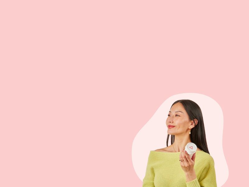 A woman wearing a lime green sweater and looking happy while holding a Wisp pill vial in front of a pink background