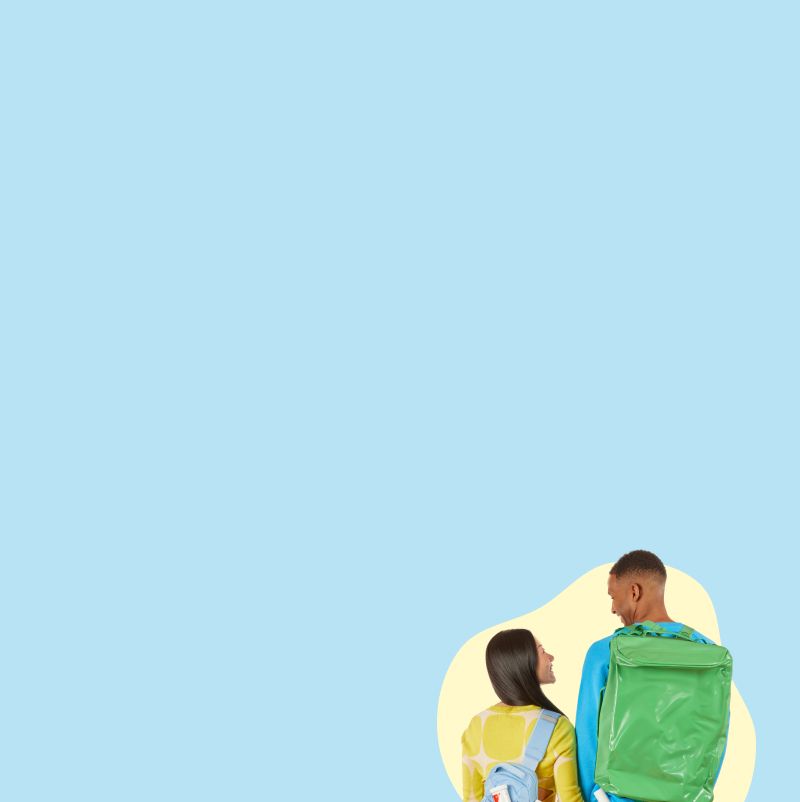 A woman wearing a yellow sweater, orange pants and a light blue backpack is smiling at and holding hands with her partner, a male wearing a blue sweater and a green backpack