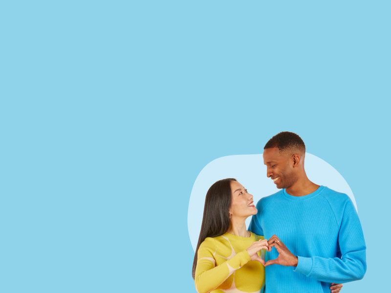 A woman and a man with they opposite hands together making a heart shape and smiling at each other in front of a light blue background