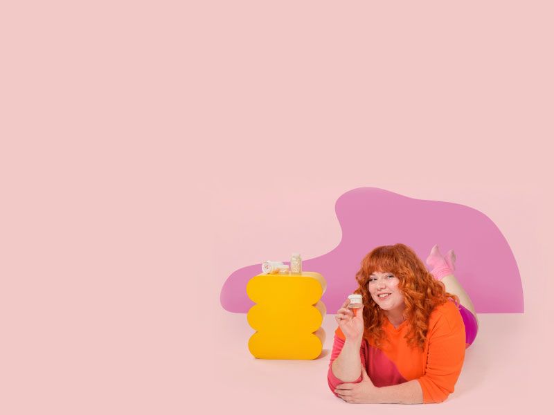 A woman wearing a red sweater and purple shorts is laying on her stomach with her feet crossed and holding a Wisp glass pill jar next to a yellow nightstand with various Wisp bottles on it