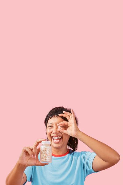A smiling female holding a Wisp glass pill jar in one hand and a a pill in the other in front of a pink background