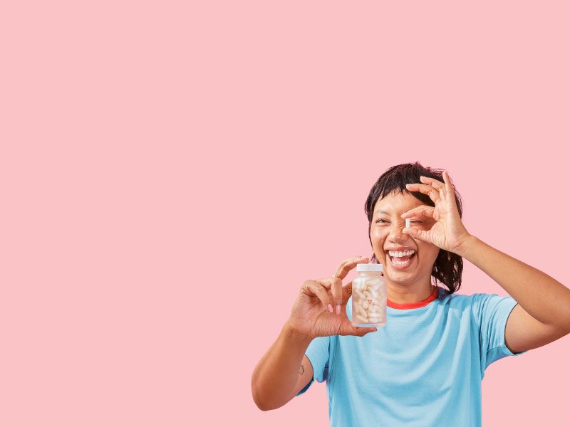 A smiling female holding a Wisp glass pill jar in one hand and a a pill in the other in front of a pink background