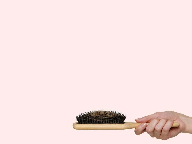 A person's hand holding a brush with excess hair in the bristles in front of a pink background