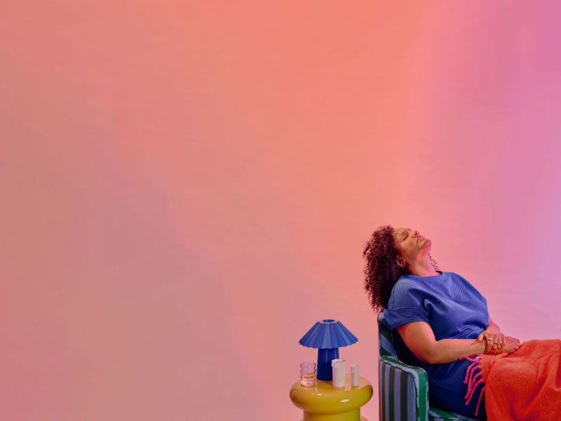 A woman wearing a blue shirt is sitting on a striped sofa with her head back in discomfort next to a yellow nightstand with a blue lamp and various Wisp medication vials