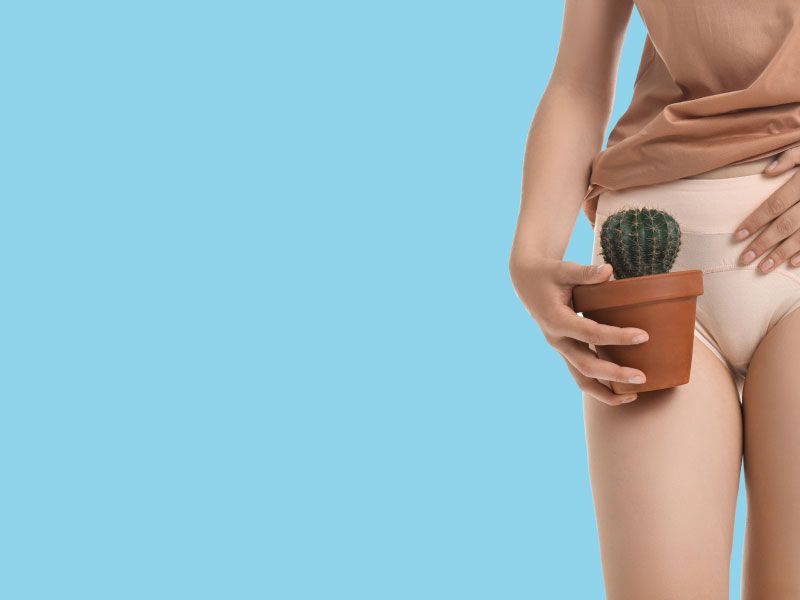 A woman standing up in white underwear and a tan shirt with a hand on her abdomen and holding a small cactus indicating pain