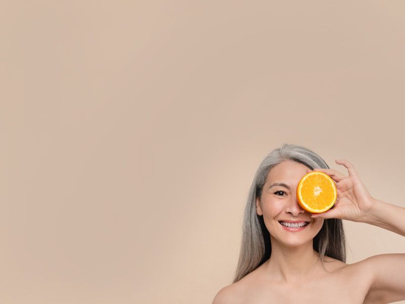 A woman is smiling and holding a sliced open orange in front of her left eye with a beige background