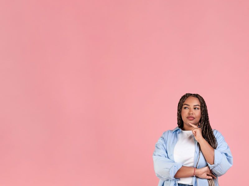 A woman wearing a white tee with a light blue button-up shirt has a finger to her chin in contemplation in front of a pink background