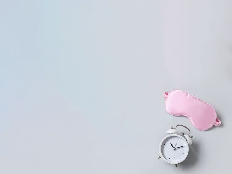 A pink sleep mask and a white alarm clock on a light gray surface