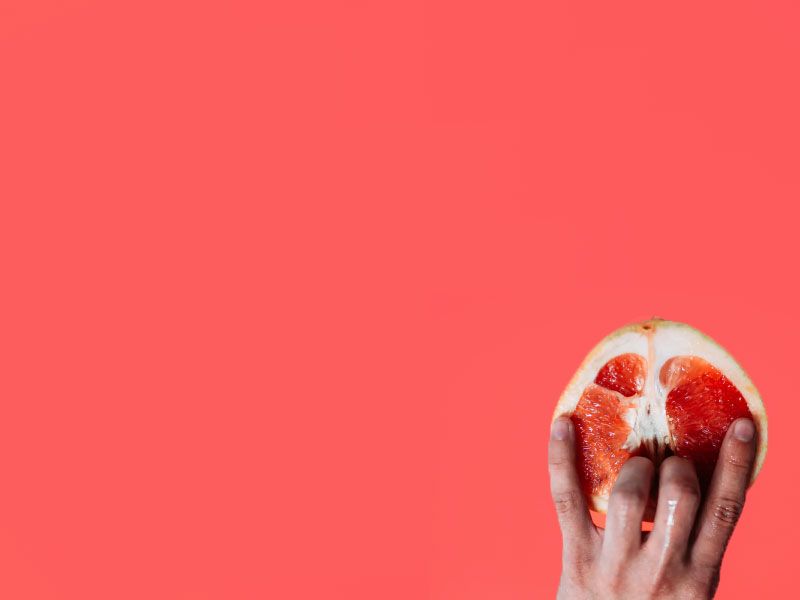 A hand suggestively holding a cut open grapefruit on a reddish background