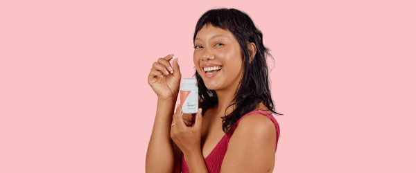 A happy woman wearing a red tank topis holding a pill and a Wisp bottle in front of a pink background
