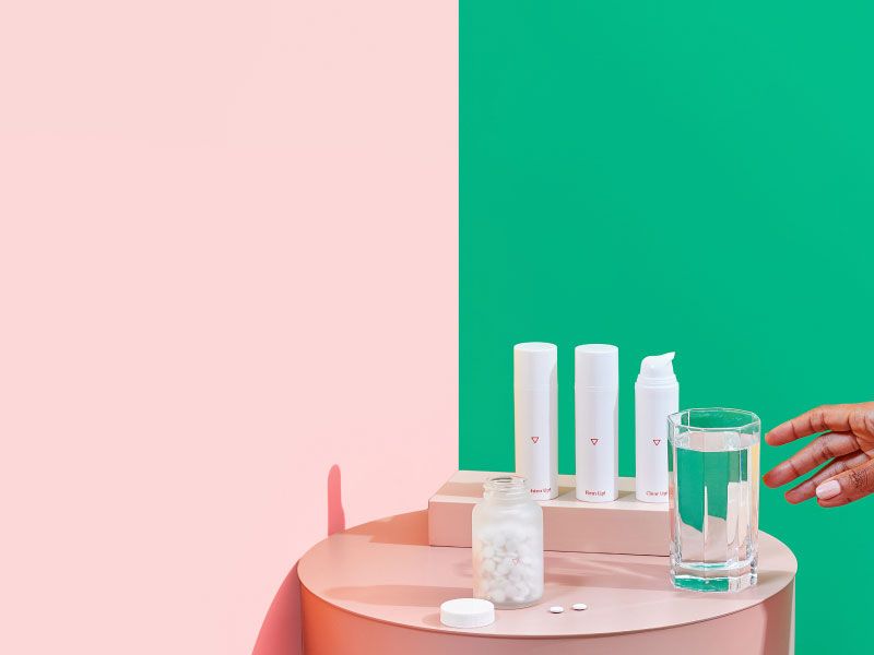 Woman's hand reaching for a glass of water on a pink nightstand with 3 bottles of Wisp Skincare treatment and a Wisp glass pill jar with pink and red bed and green and pink walls