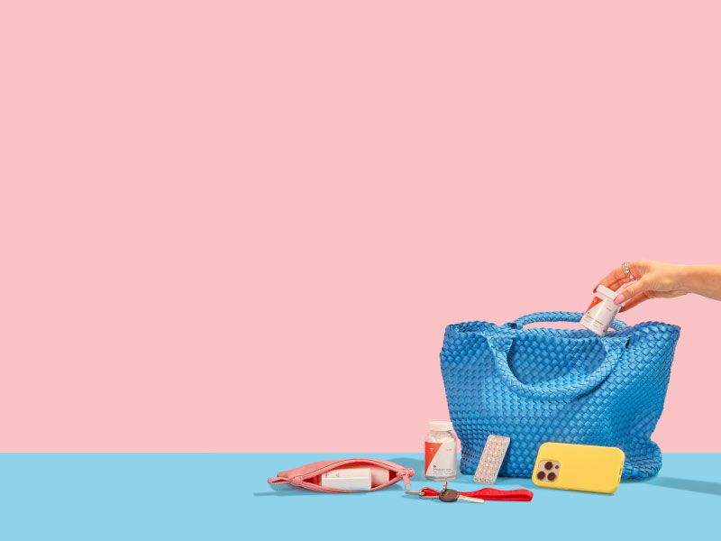 A person's hand is putting Wisp Equalizing Probiotics into a blue handbag with other Wisp Medication, a mobile phone, keys and a travel pouch on a blue surface with a pink background