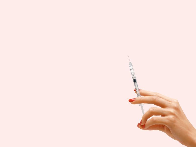 A woman's hand holding a syringe in front of a light pink background