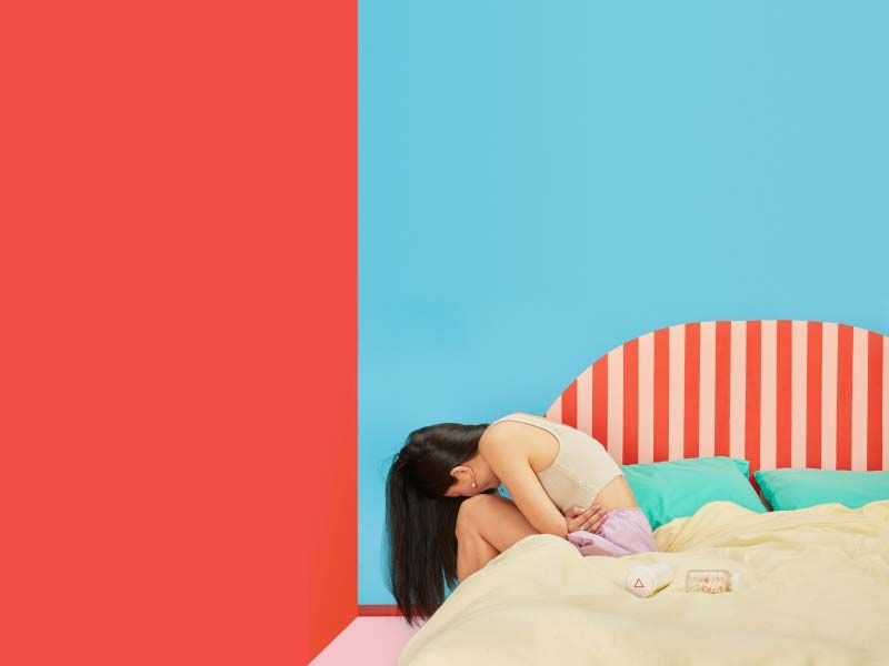A female sitting on a bed doubled over in apparent discomfort with medication on the bed next to her